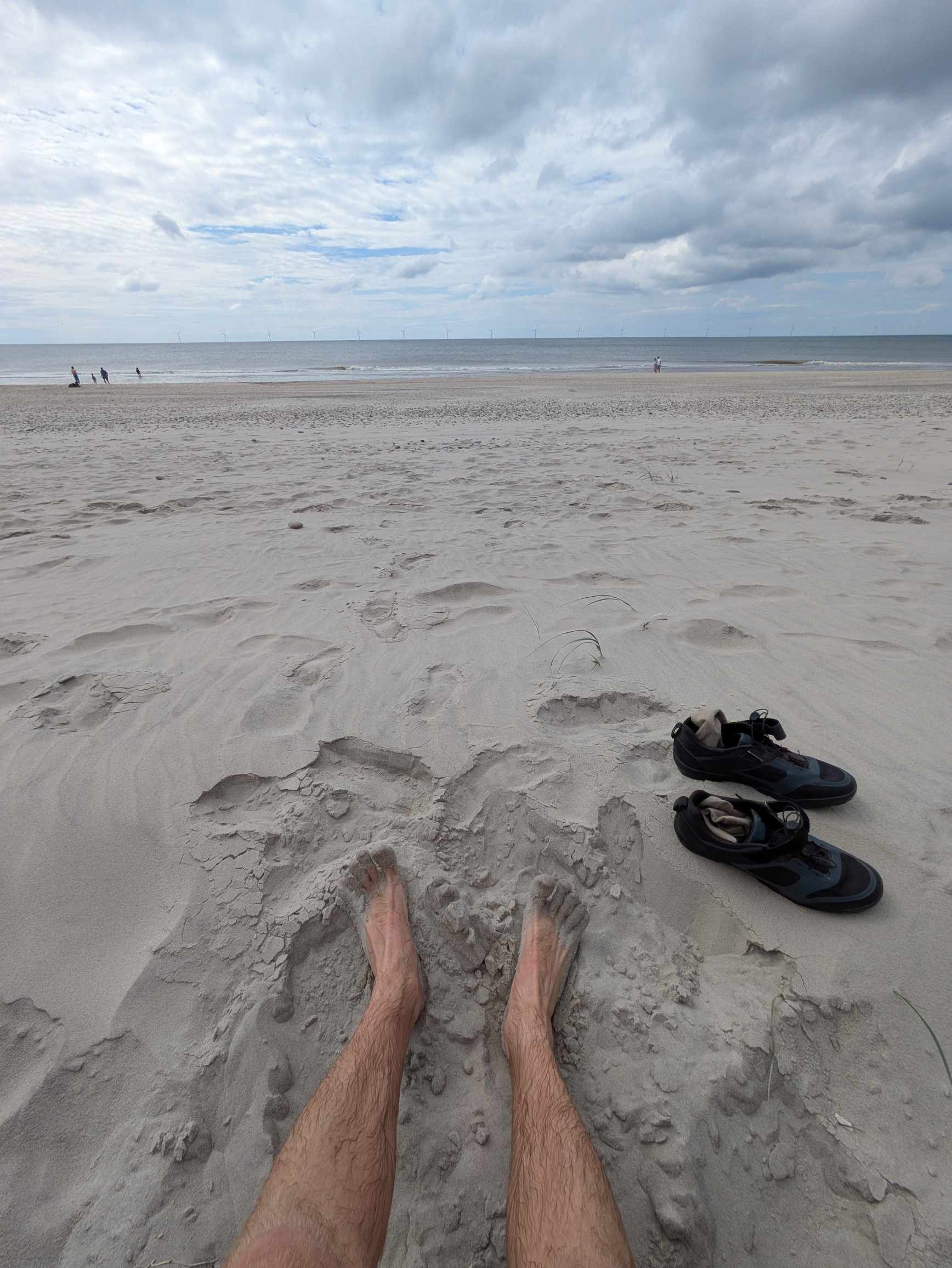 Bloße Beine liegen auf einem Sandstrand in Richtung Meer; rechts liegen schwarze Sandalen im Sand.