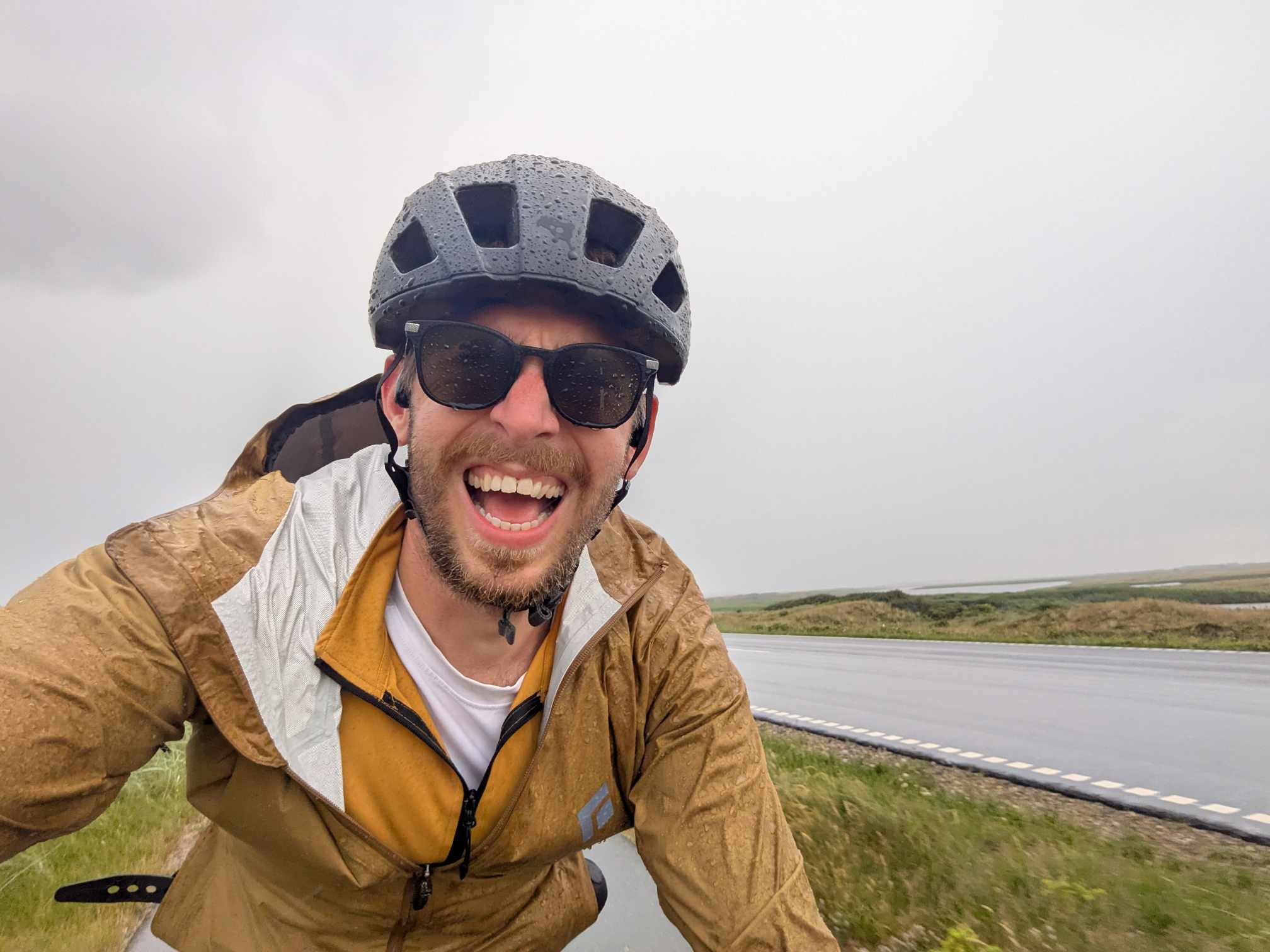 Lächelnder Radfahrer mit Helm, dunkler Sonnenbrille und hellbrauner Jacke neben einer asphaltierten Straße.