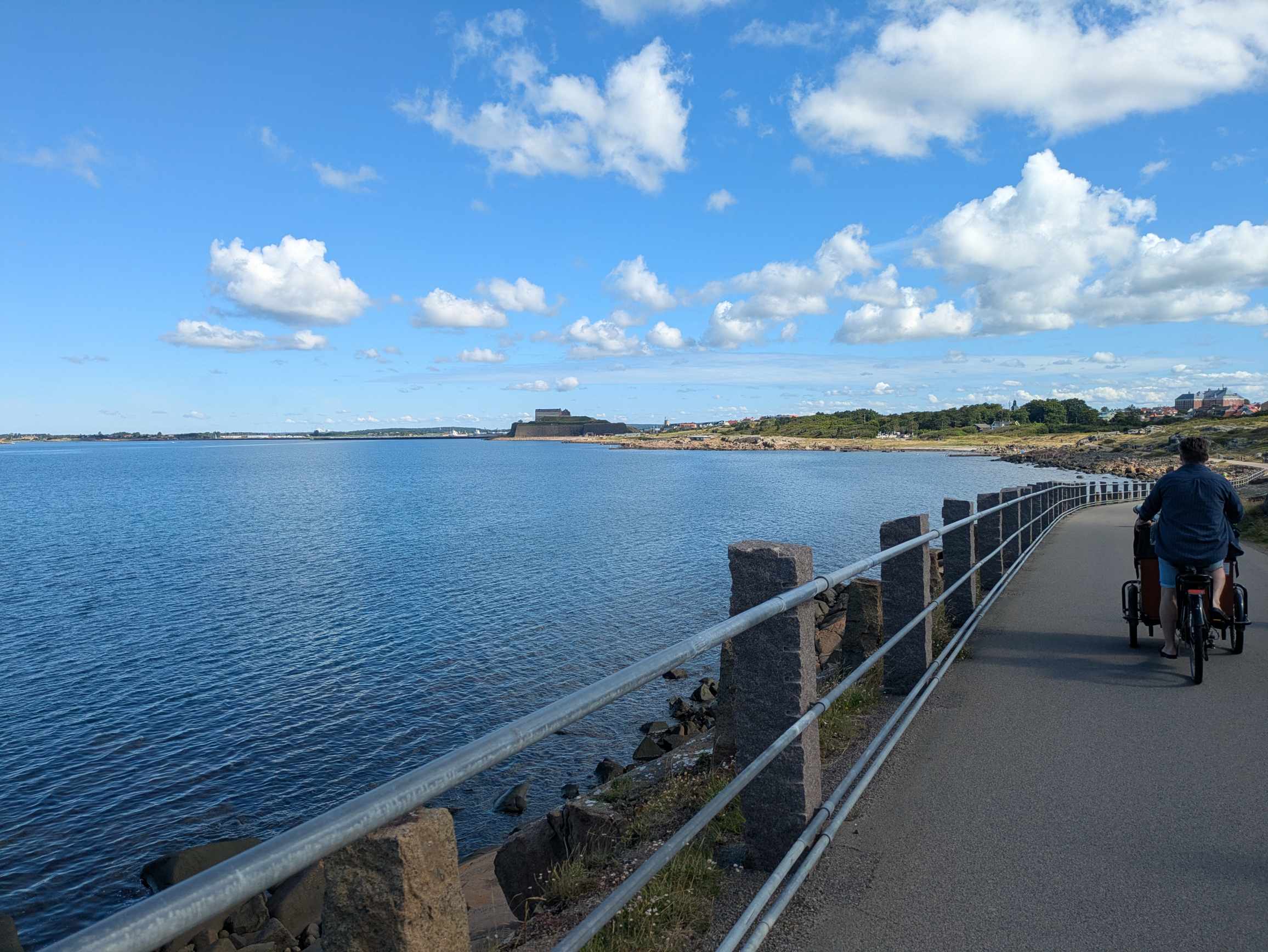 A cyclist on a lakeside path with a railing next to blue water; in the background, a flat stone structure, buildings, and clouds.