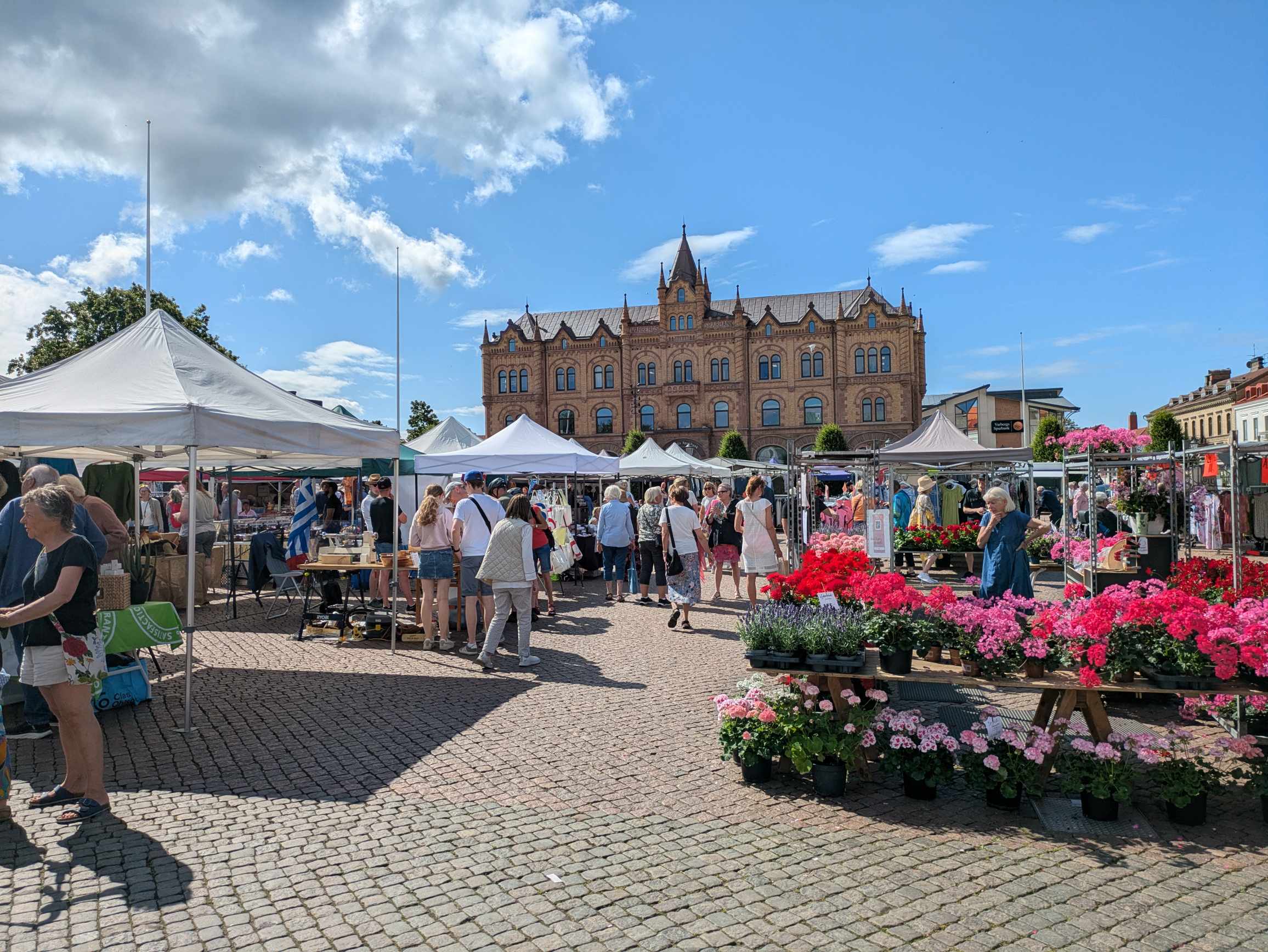 A bustling market on a cobblestone square with white market tents, flowering potted plants, and a historic brick building.