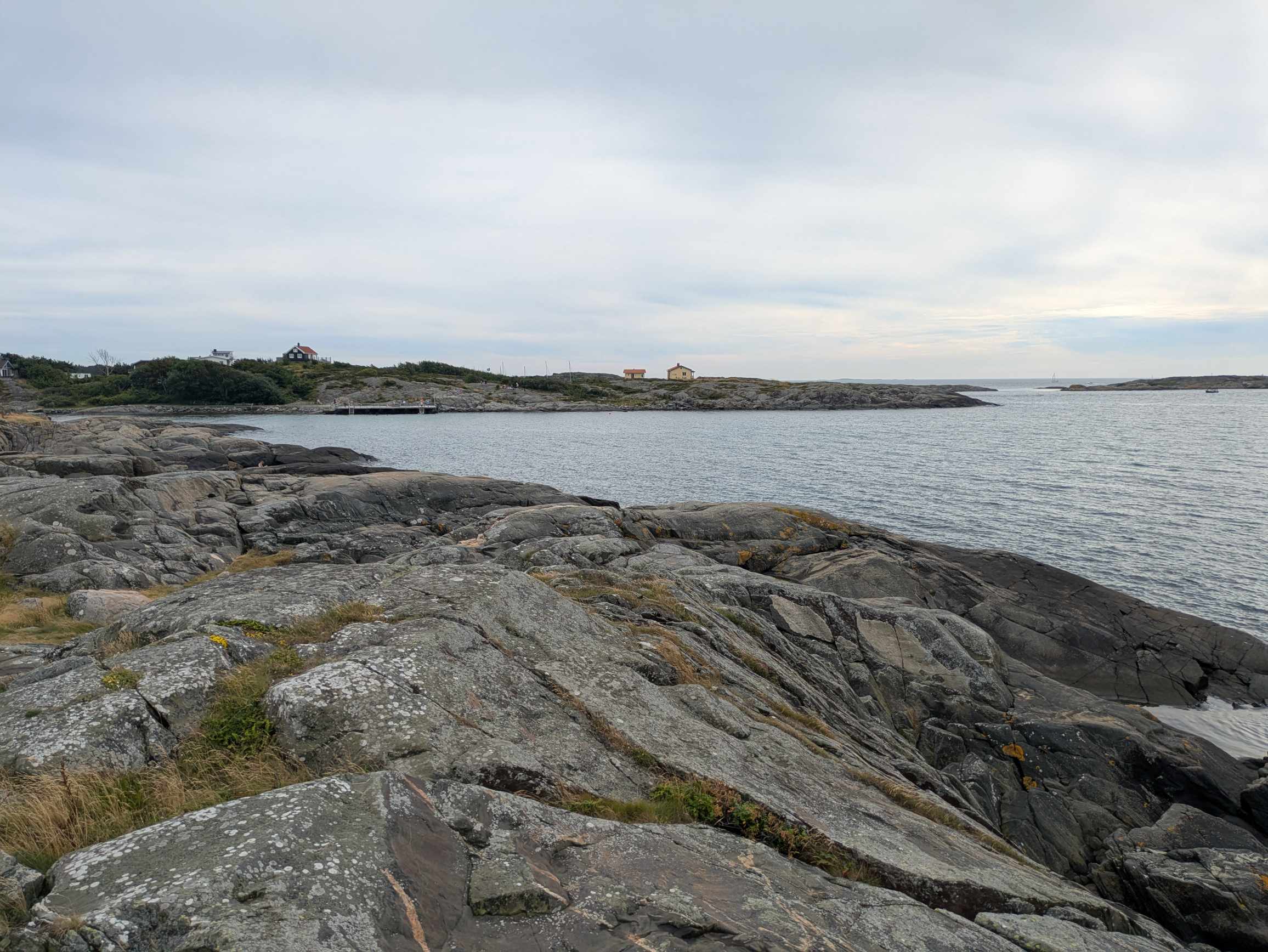 A rocky coastline with calm waters, small houses, a pier, boats in the distance, and flat islands under an overcast sky.