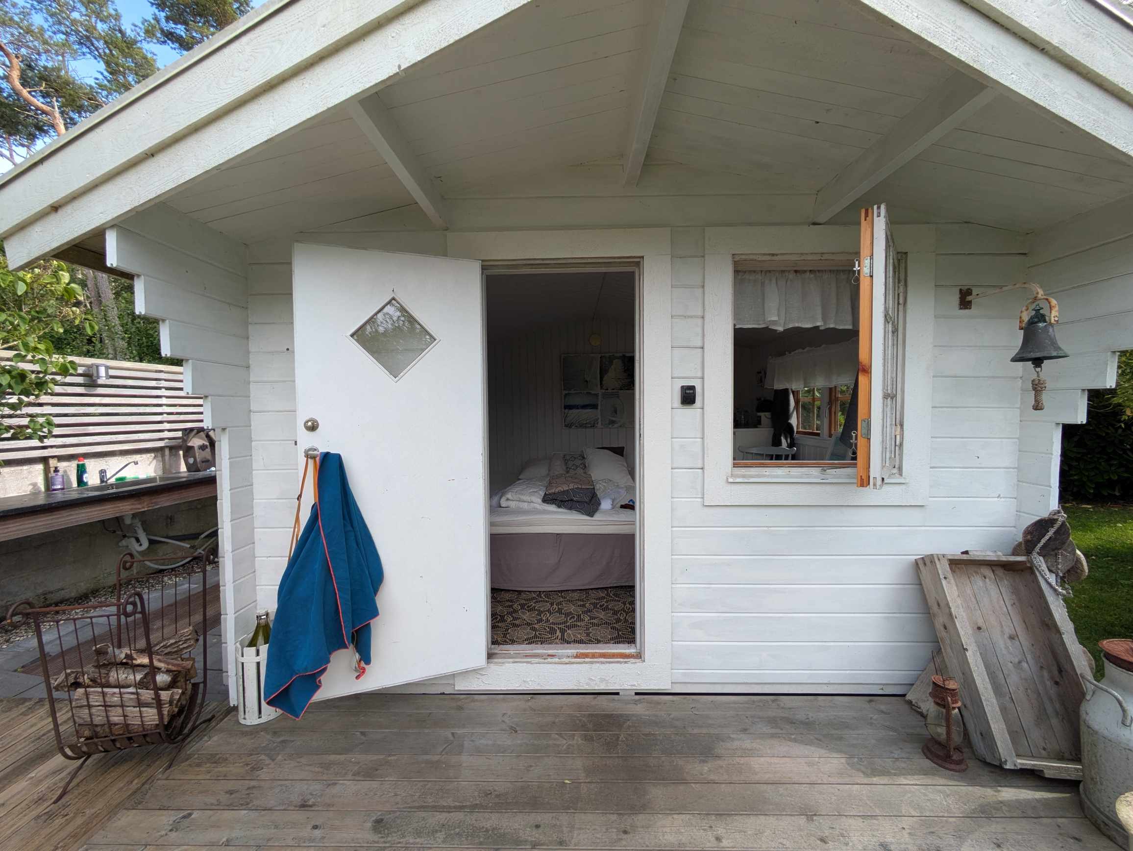A white wooden cabin on a terrace with an open door leading to the bed, a window with a shutter flipped up, an outdoor sink, and a hanging bell.